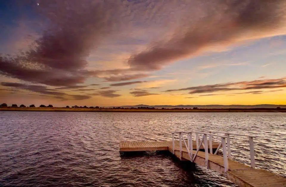 Sunset over a calm lake with a wooden dock extending into the water and colorful orange and purple clouds in the sky