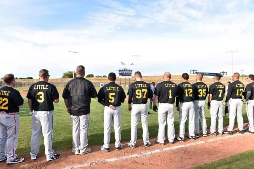 Baseball players stand lined up along the infield dirt backs to the camera wearing black jerseys with LITTLE and yellow numbers