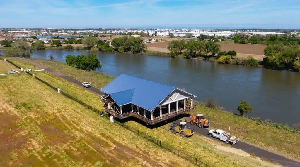 RIBlogImagesJuly2019WelcomeCenter1 1 River Islands Aerial view of a blue roofed pavilion on a riverbank with construction trucks and a parked white vehicle nearby