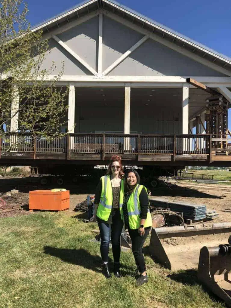 Two women in high visibility vests smile at the camera in front of a large elevated building under construction on a sunny day
