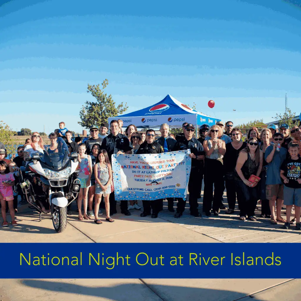 Community group including police officers and families posing with a National Night Out banner at River Islands, with a police motorcycle and Pepsi tent in the background.