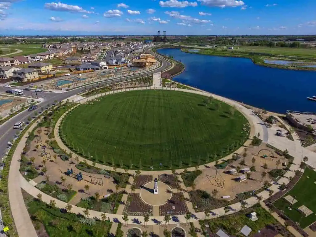 RIParkMichaelVegaPark1 River Islands Aerial view of a circular park with a large central green field surrounding walking paths and a lake to the right beyond housing areas