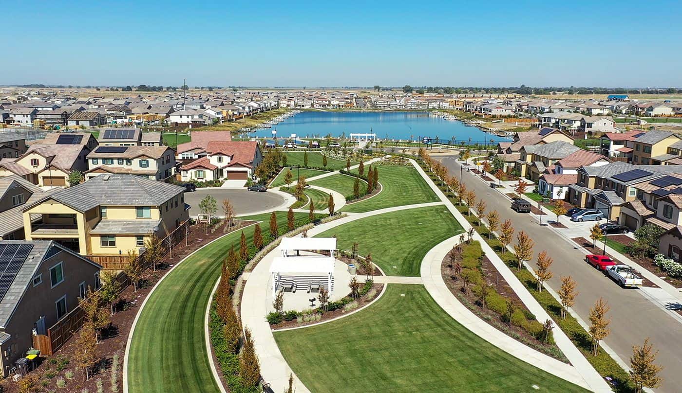 Aerial view of a suburban park with winding concrete paths lush green lawns and a lake bordered by houses in the distance