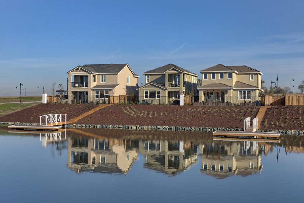 Row of beige suburban houses along a waterfront with wooden docks and a clear blue sky, reflected in the calm water.