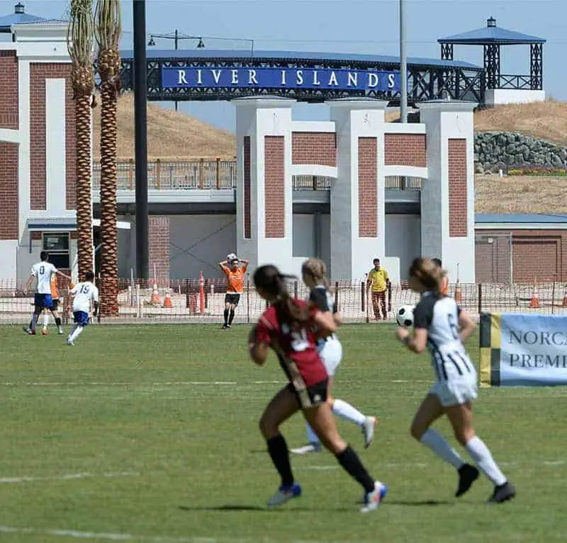 2019photo2 River Islands Youth girls soccer match on a sunny day at River Islands stadium players in red and white chase the ball on a grassy field with brick arches and spectators in the background