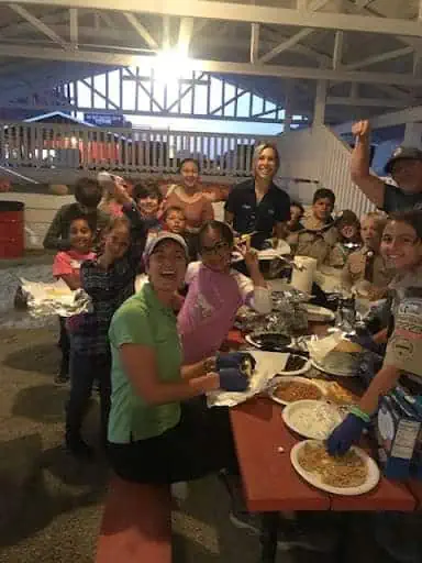 2019photo6 River Islands Group of smiling volunteers and children share a meal at a long outdoor table during a community dinner event with pasta plates and beverage cups visible