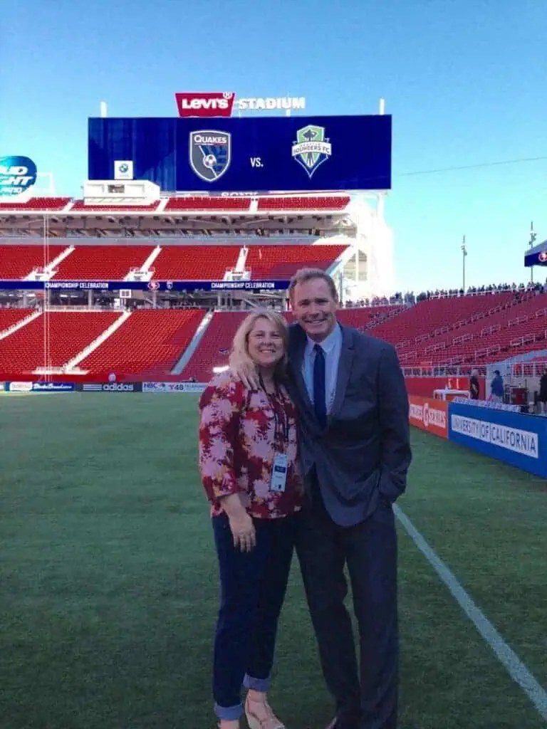 7809483426000630234089175152842272701677568o River Islands Smiling couple in smart casual attire posing on a soccer pitch at Levis Stadium with a large scoreboard behind them showing Quakes vs Founders FC and red seating around the stands