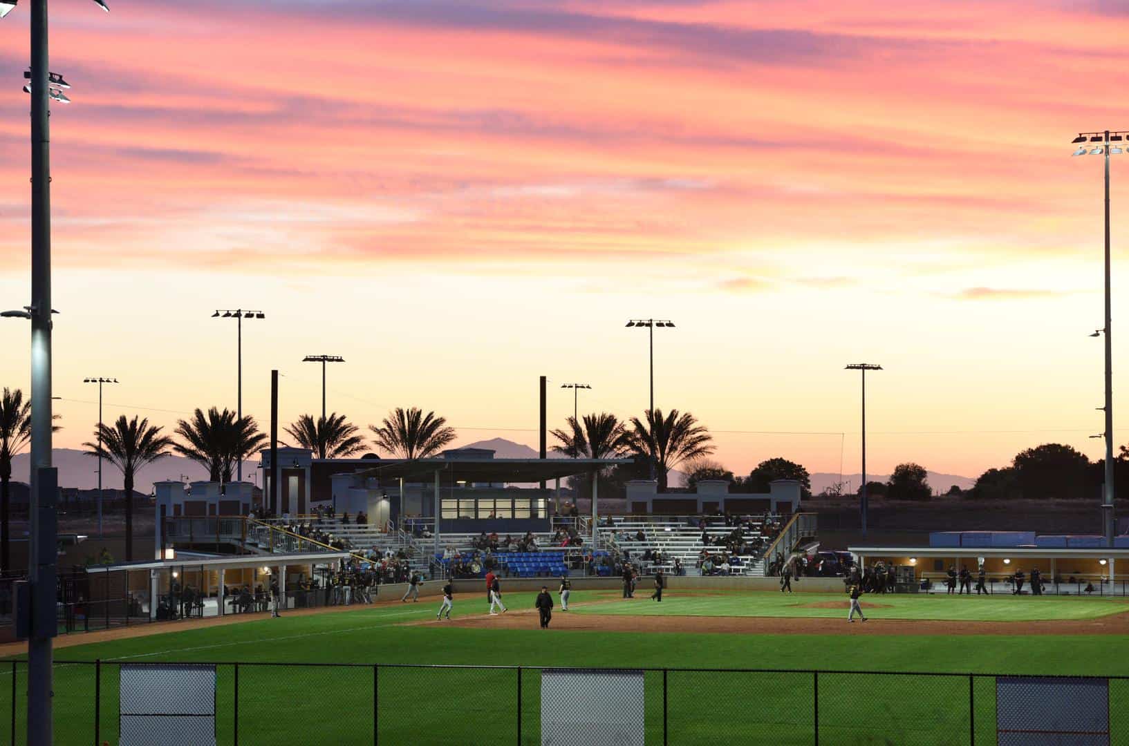 Baseball field at sunset with players on the infield and spectators in bleachers under a pink orange sky and tall light poles in the background