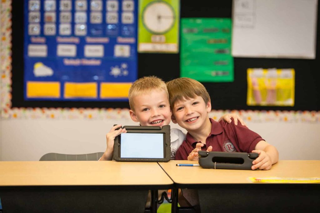 Two smiling young students sit at a classroom desk, each holding a tablet in a protective case and looking at the camera. Background shows a colorful bulletin board and classroom posters.