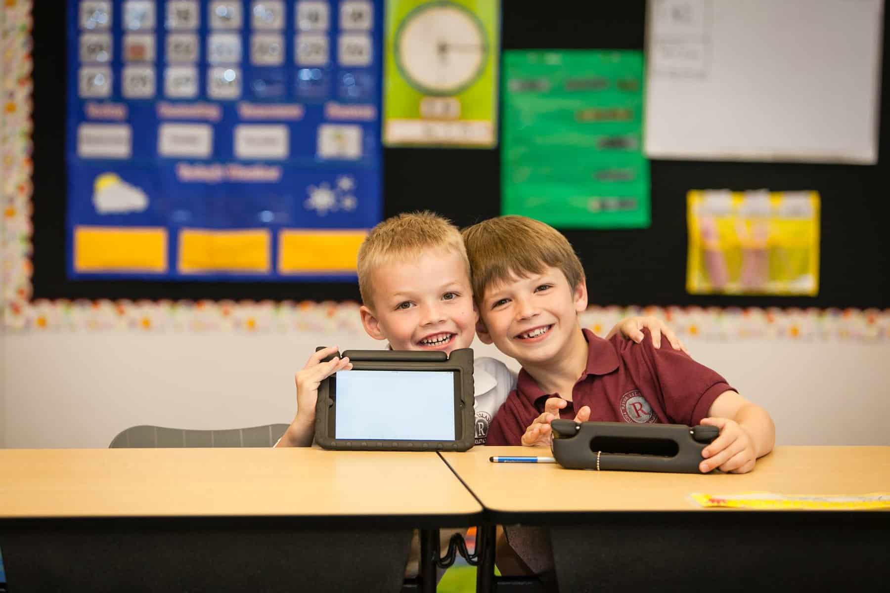 Two smiling young students sit at a classroom desk each holding a tablet in a protective case and looking at the camera Background shows a colorful bulletin board and classroom posters