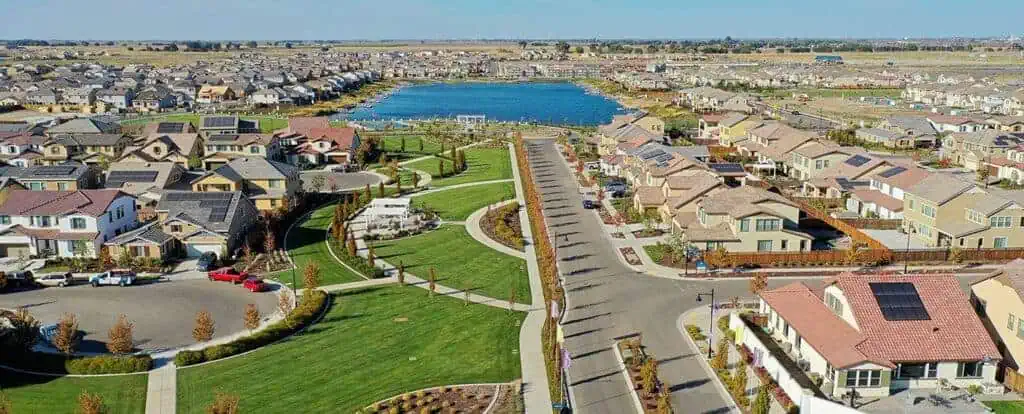 Aerial view of a suburban neighborhood with a central green park, winding paths, and a lake in the distance.
