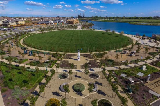 Aerial view of a circular park with a large green lawn central monument and surrounding pathways beside a blue lake