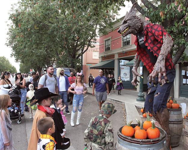Tall werewolf figure in a plaid shirt towers over pumpkins while a crowd watches at a street festival by a tree-lined sidewalk.