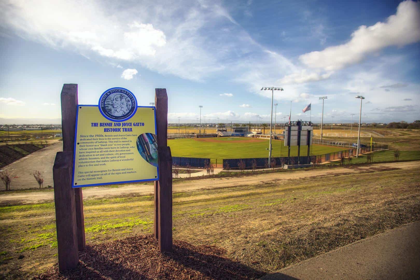 Signboard for the Bennie and Joyce Gatto Historic Trail beside a baseball field under a blue sky with scattered clouds in a park