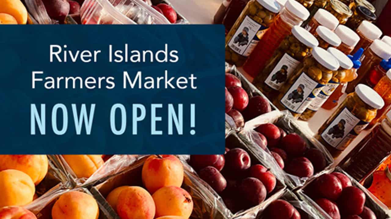 Blue sign reading River Islands Farmers Market NOW OPEN over fruit and bottled goods at a market stall
