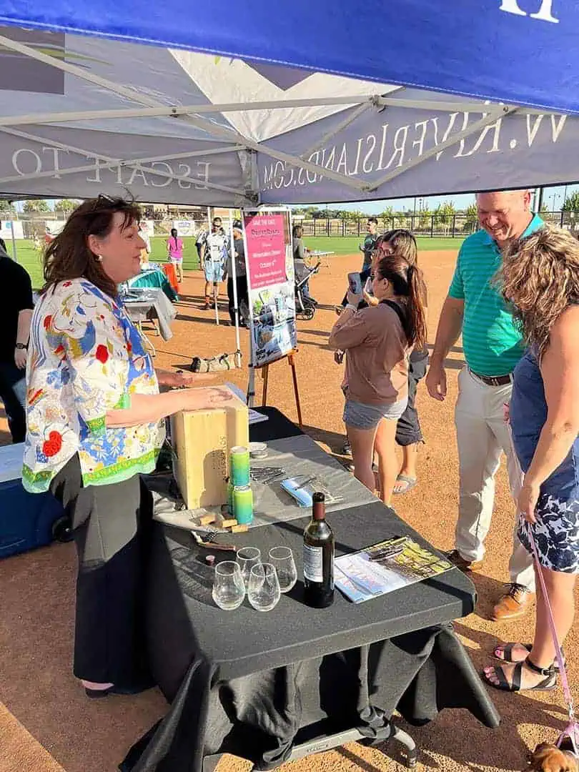 RINNOBlogPhoto4 River Islands Informational booth at an outdoor event on a baseball field a woman stands behind a table with a wine bottle several glasses and flyers as attendees mingle nearby