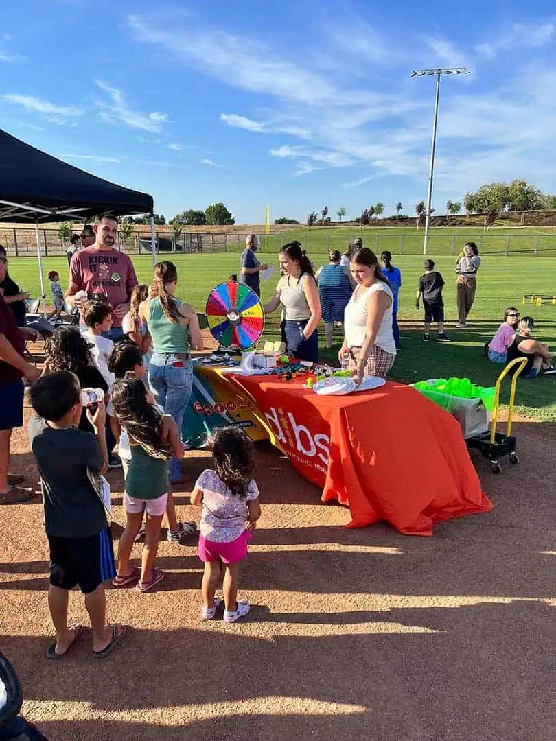 RINNOBlogPhoto5 River Islands Group of kids and adults at an outdoor community fair around a prize wheel on a table under a canopy on a sunny day at a sports field