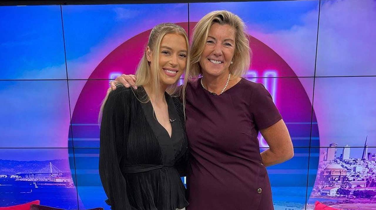 Two women pose together in a TV studio smiling at the camera with a colorful set behind them