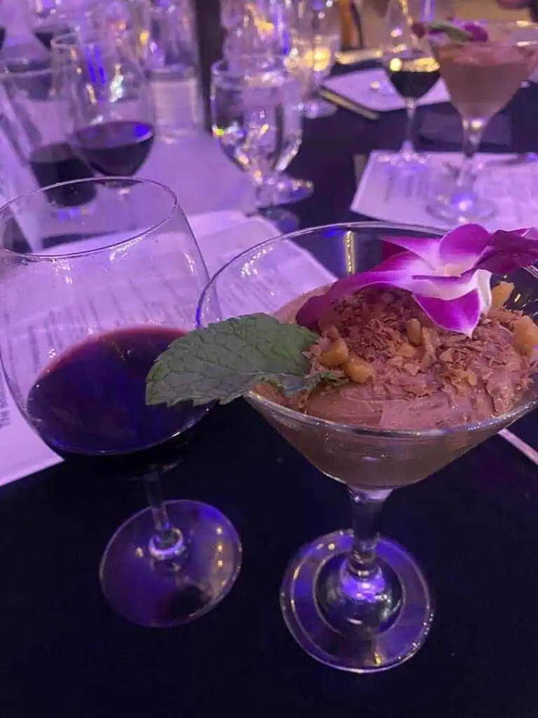5IMG8687geo River Islands Coupe glass cocktail with a flower garnish crushed nuts topping and a mint leaf on a dark banquet table with purple lighting and wine glasses in the background