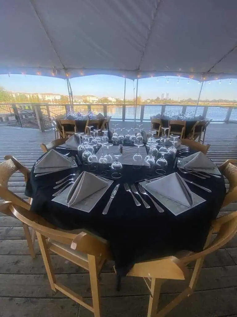 8IMG8655geo River Islands Round table set for a formal outdoor dinner on a wooden deck under a canopy with black tablecloth folded napkins and glassware by the water edge