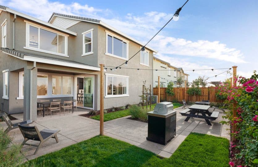 Backyard of a two story beige house featuring a concrete patio outdoor dining table lounge chairs a grill and string lights overhead
