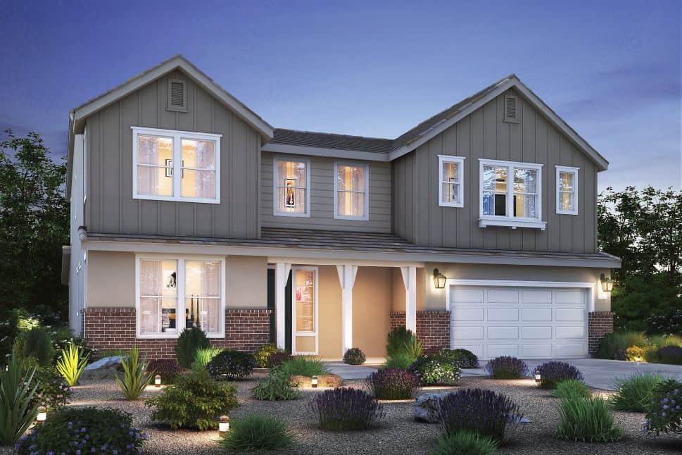 Two story suburban house at dusk with gray siding brick accents illuminated windows and a two car garage