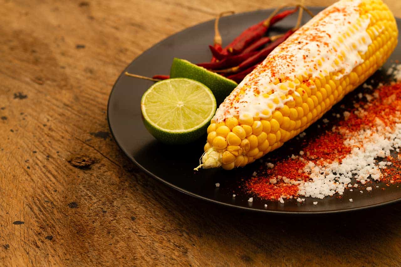 Corn on the cob served on a black plate with lime half red chilies and a dusting of salt and chili powder on a rustic wooden table