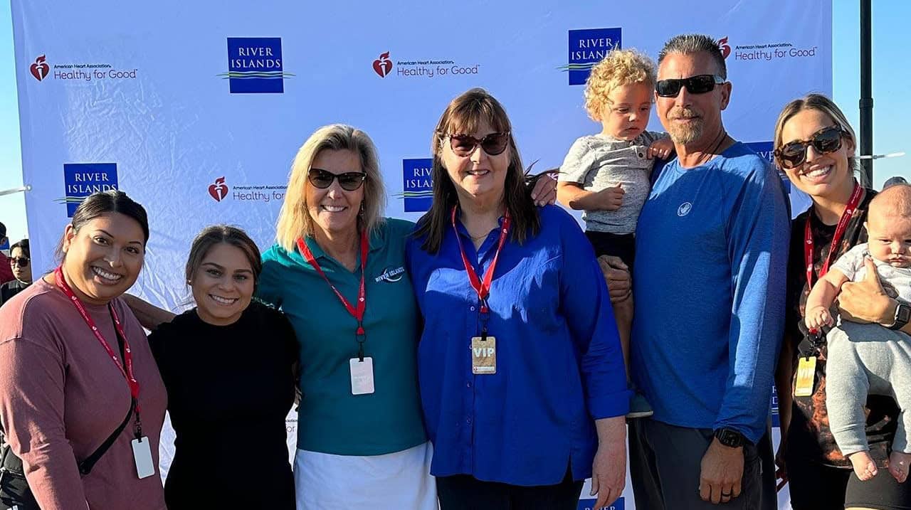 Group of six adults and two children posing for a photo in front of a blue River Islands backdrop at an outdoor event Lanyards and badges are visible on several people