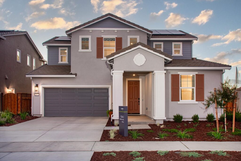 Two story gray suburban house with a two car garage white columns and brown front door solar panels on the roof and landscaped front yard