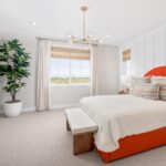 Spacious primary bedroom in Bridgeway Plan 3502 with modern chandelier, large windows, and upholstered orange bed.