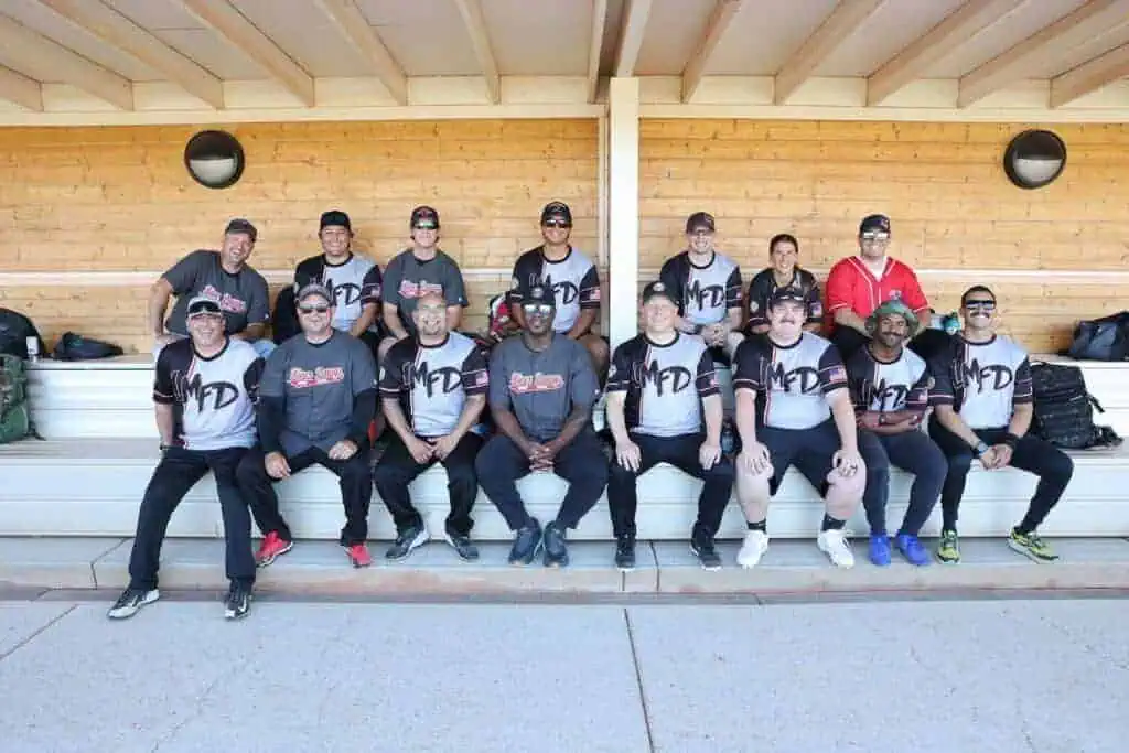 Group of 14 athletes in matching gray and black MFD jerseys posing on bleachers inside a wooden dugout