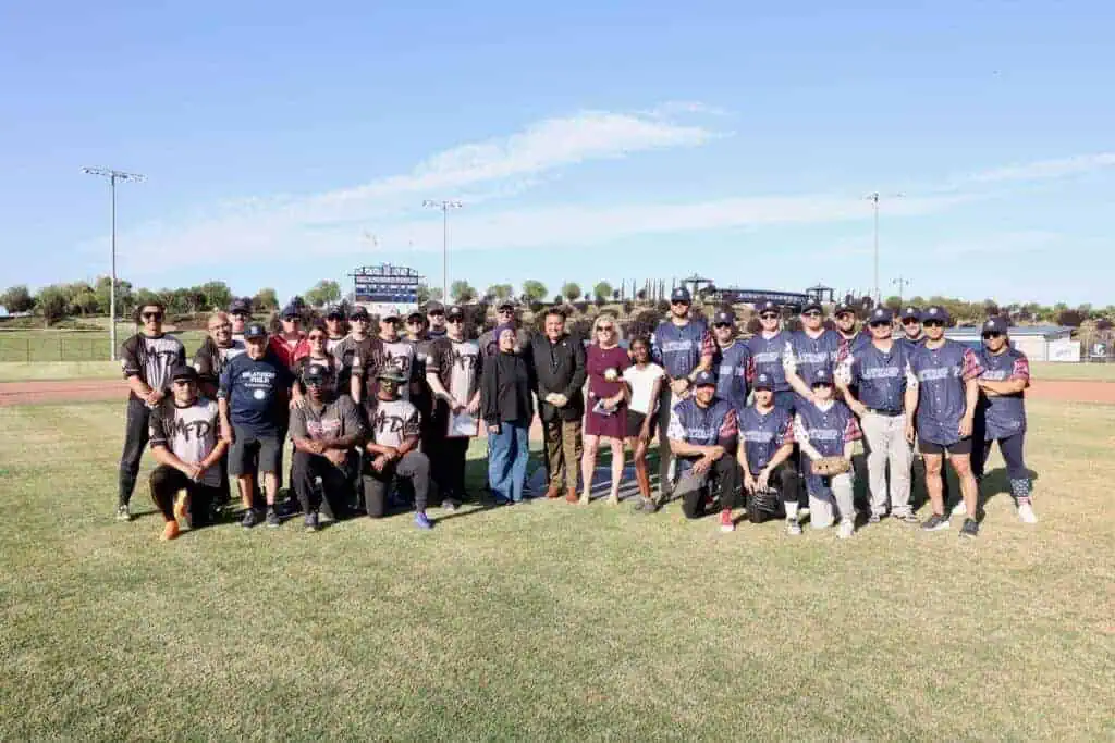Large group of people in baseball uniforms posing for a team photo on a sunny baseball field with a scoreboard in the background