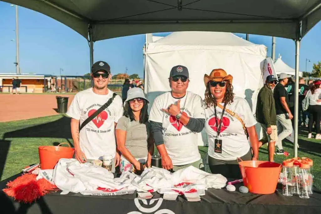 HW2519geo River Islands Four volunteers stand behind a table at an outdoor charity event under a canopy wearing white shirts with a red heart logo and smiling at the camera