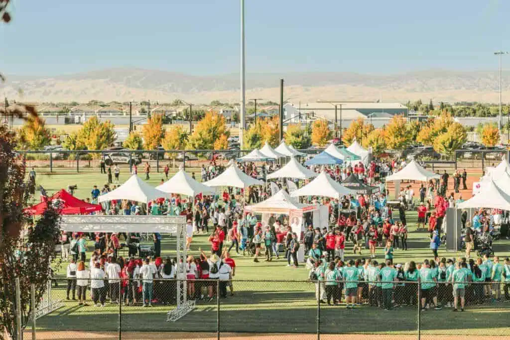 HW2524geo River Islands Crowd at an outdoor fair on a sports field with white tents booths and fall colored trees in the background identifying the event setting at a park like venue
