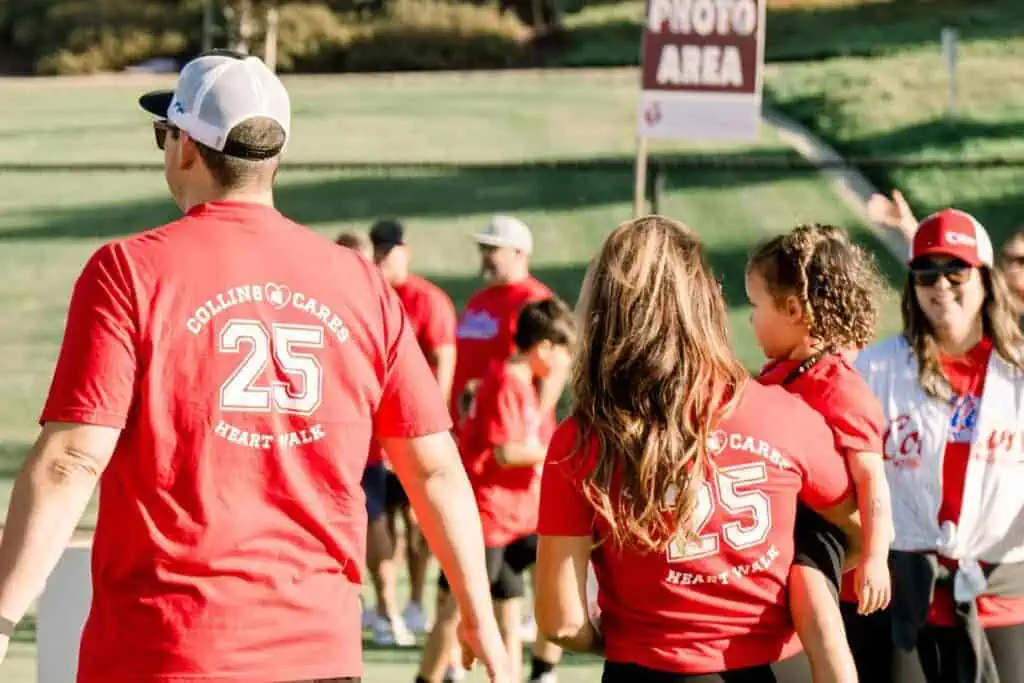 HW2534geo River Islands Back view of a man in a red Heart Walk shirt with the number 25 walking with a woman and child at an outdoor event a Photo Area sign is in the background