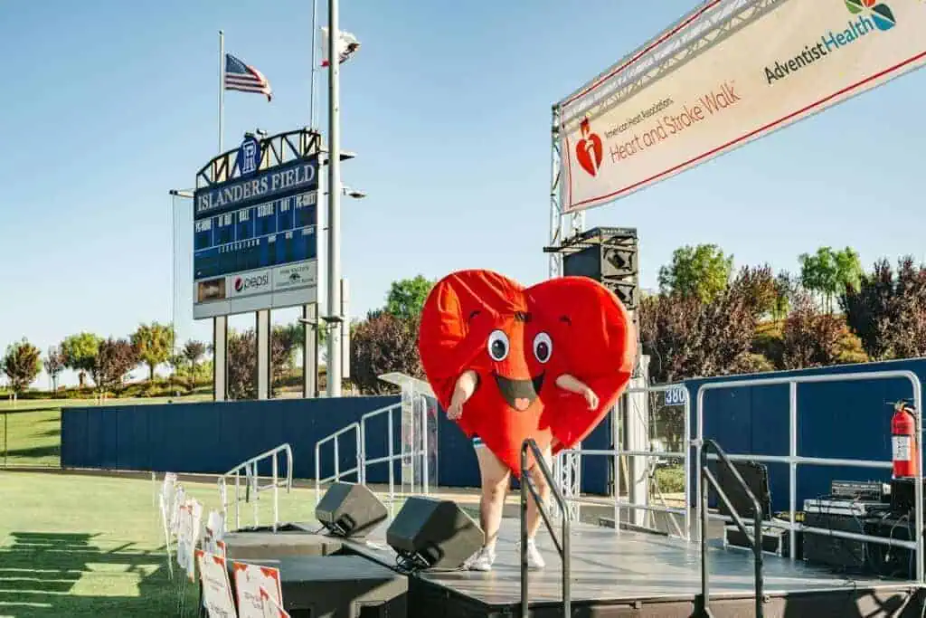 HW2542geo River Islands Red heart mascot dancing on a stage at a Heart Walk event outdoors with banners and scoreboard in the background at Islanders Field