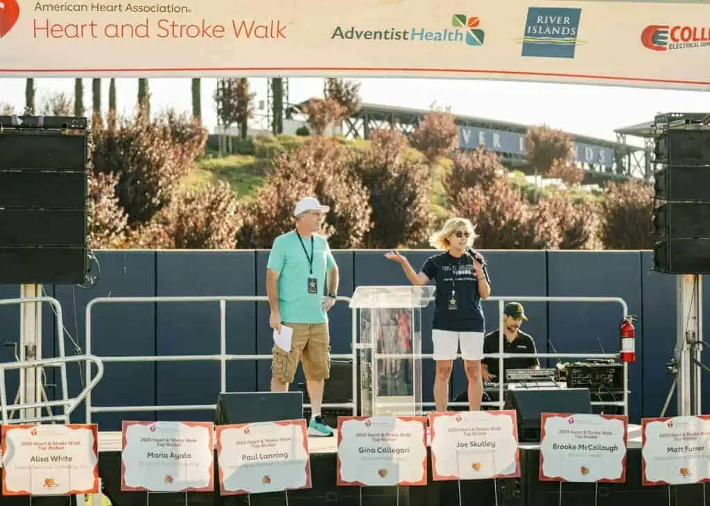 HW2550geo River Islands Stage setup for a Heart and Stroke Walk event with two hosts at a clear podium and sponsor banners overhead on a sunny day names displayed on front boards nearby