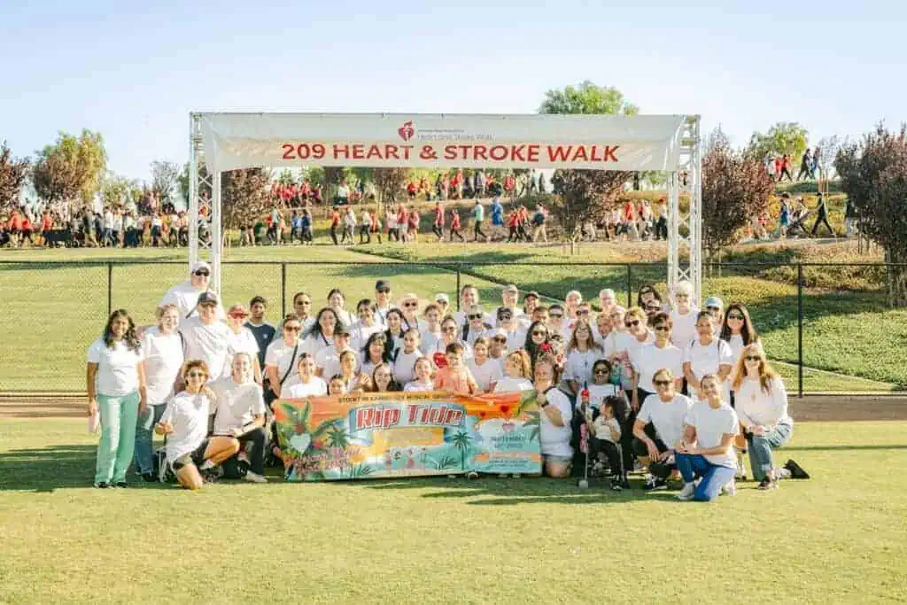 HW2564geo River Islands Large group of volunteers in white shirts pose on a sports field under a banner reading 209 Heart Stroke Walk with a colorful Rip Tide banner in front
