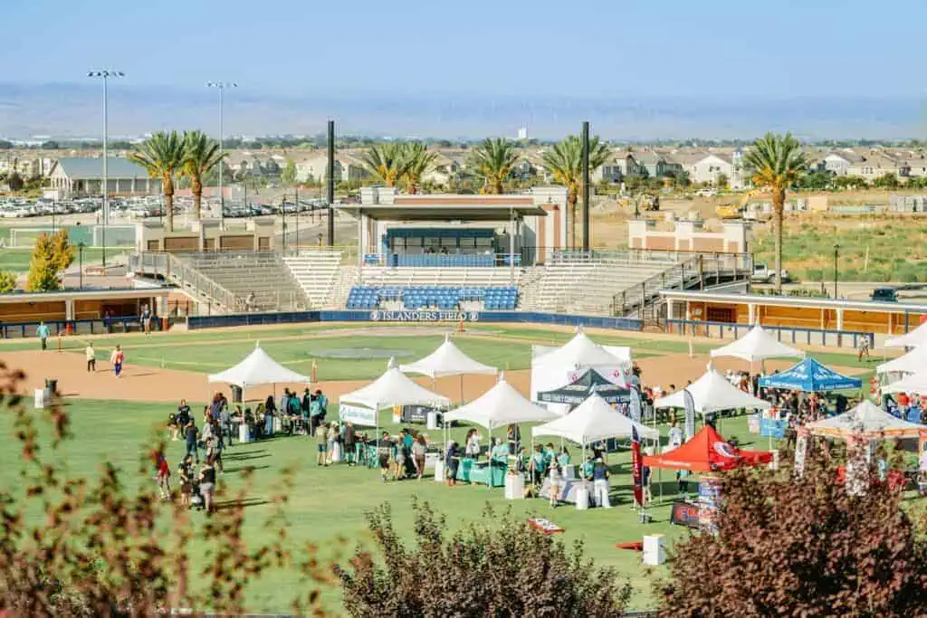 Community event at a baseball field with white tents and people browsing stalls on a sunny day on the field edge of the stands