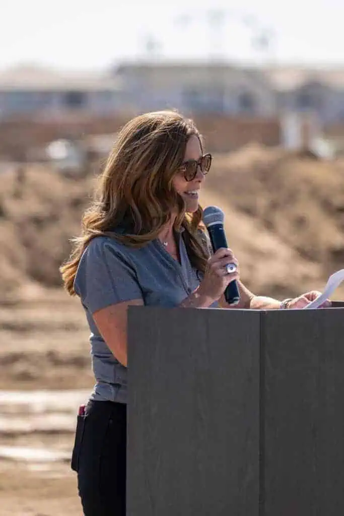 IMG0618geo River Islands Woman speaking into a microphone at a wooden podium outdoors holding papers with a blurred construction site background