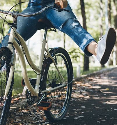 Person performing a wheelie on a beige bicycle along a wooded path one leg kicked out to the side