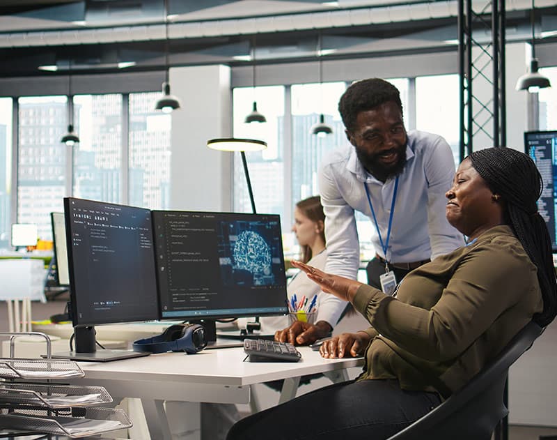 Two colleagues collaborate at a desk with dual monitors displaying code and a brain image in a bright open plan office