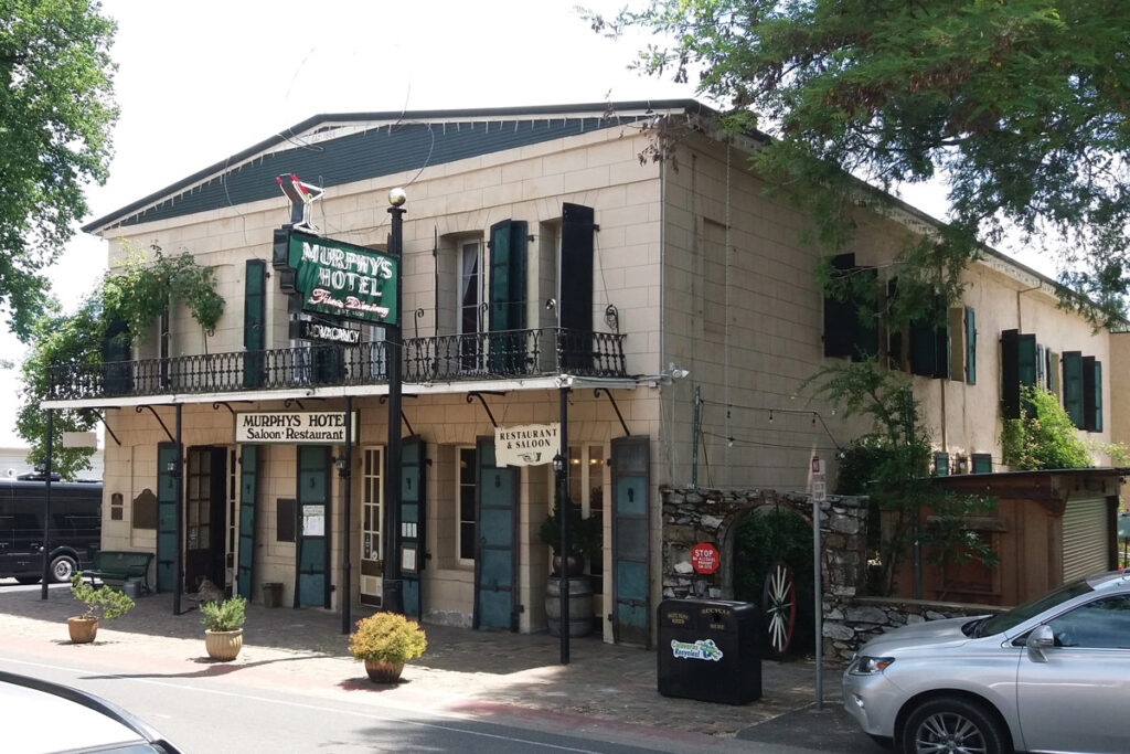 Historic two story building labeled Murphys Hotel with a hanging sign and green shutters street front with potted plants and balcony railing