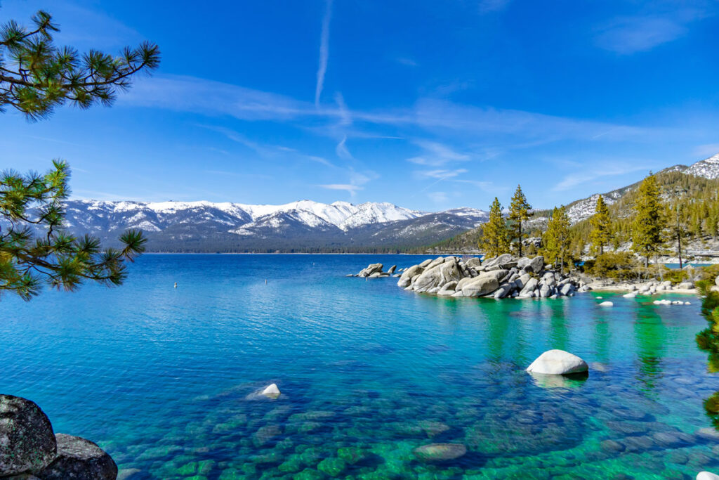 Crystal clear alpine lake with turquoise water rocky shoreline and pine trees snow capped mountains in the background under a blue sky