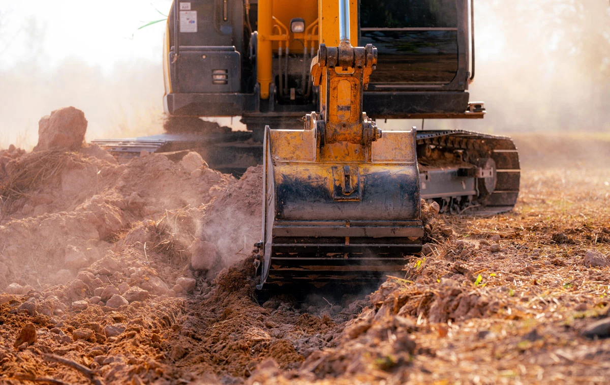 Yellow excavator with a digging bucket scooping dirt on a construction site creating a dusty trench nearby