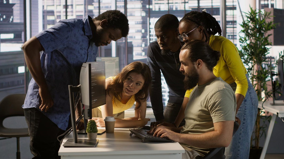 Five colleagues gathered around a computer monitor collaborating at a bright office desk