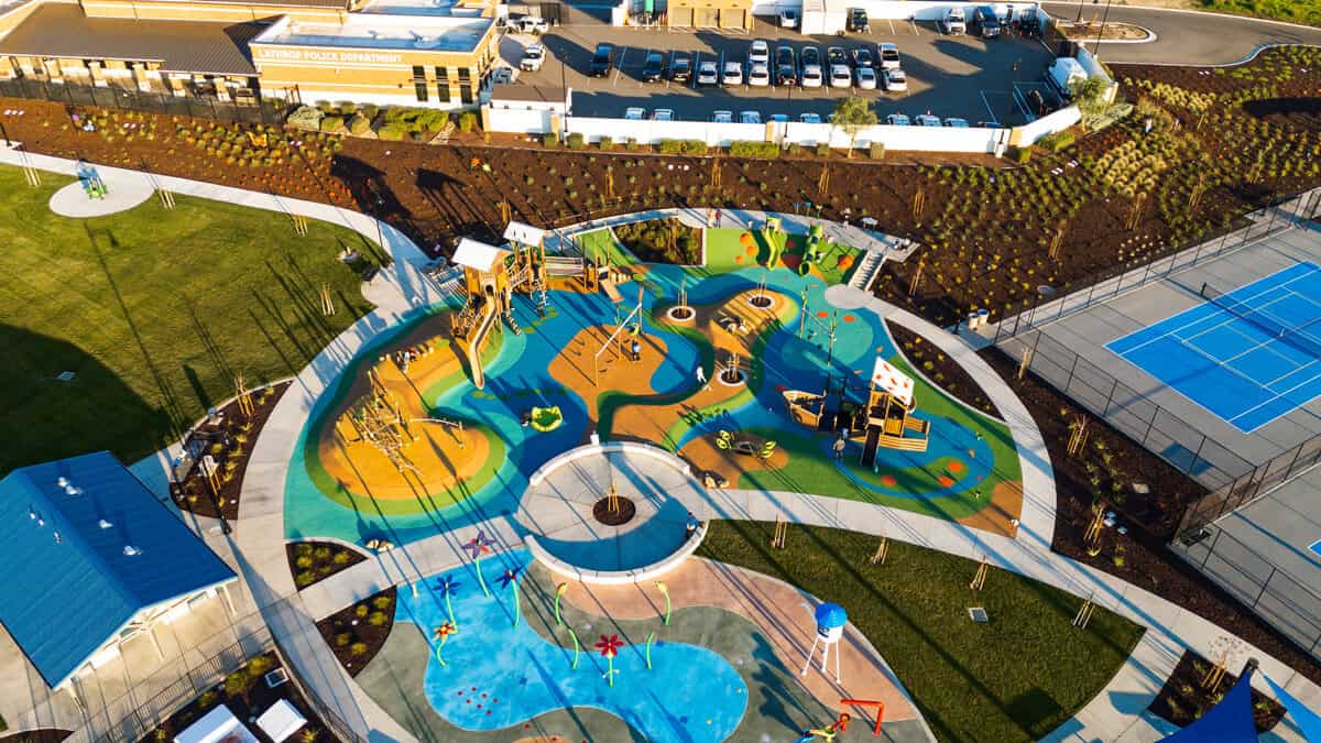 Aerial view of a large colorful playground with winding paths and water features surrounded by grass and a basketballtennis area nearby