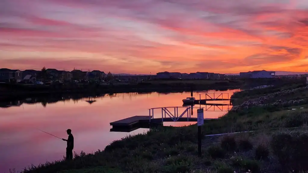 Silhouetted person fishing along a calm river at sunset with a dock and houses reflected in the water