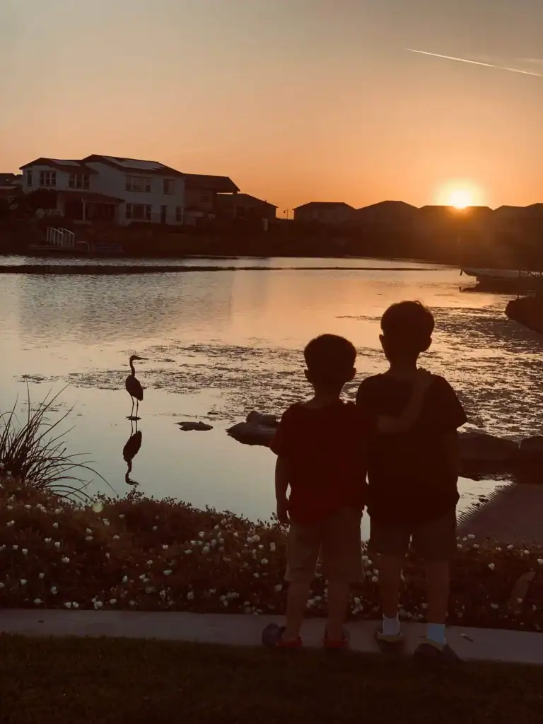 Two silhouetted children stand by a pond at sunset with a heron reflected in the water and houses in the background