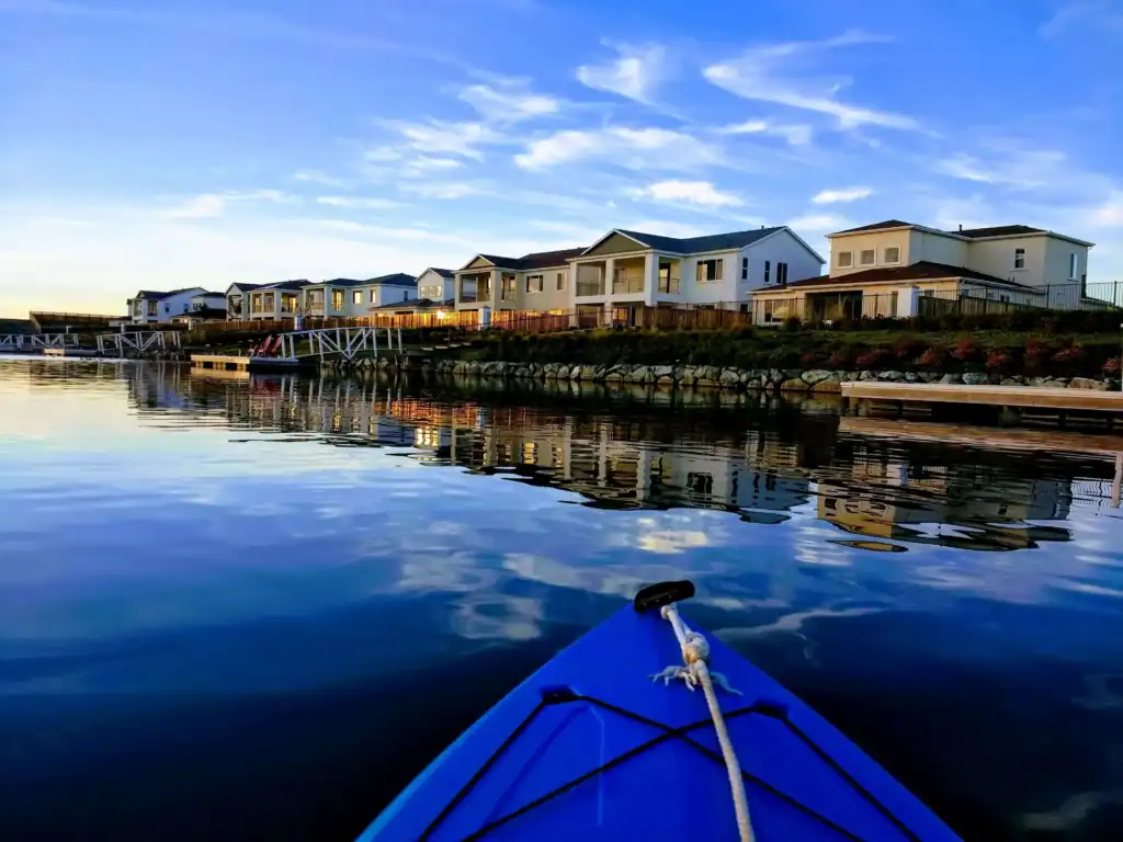 Blue boat bow on calm water reflecting a row of white waterfront houses with docks and a stone embankment
